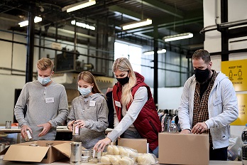 Volunteers wearing masks pack food into boxes in a warehouse setting.