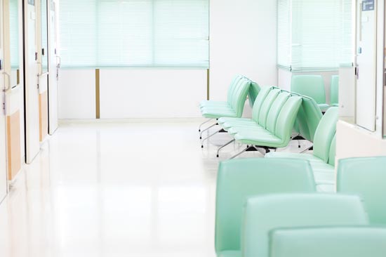Empty hospital waiting room with rows of green chairs and large windows with blinds.