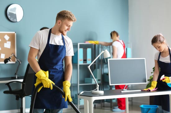 Office cleaning team vacuuming, dusting, and organizing an office space with modern furniture and blue walls.