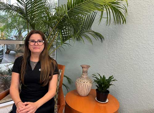 Woman sitting in a chair indoors near a small table with a potted plant and vase, with large green plant in background.
