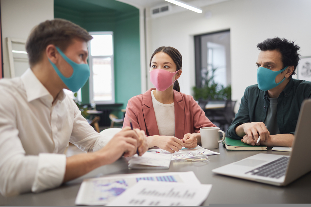 Three professionals in face masks discussing at a conference table with laptops and papers.