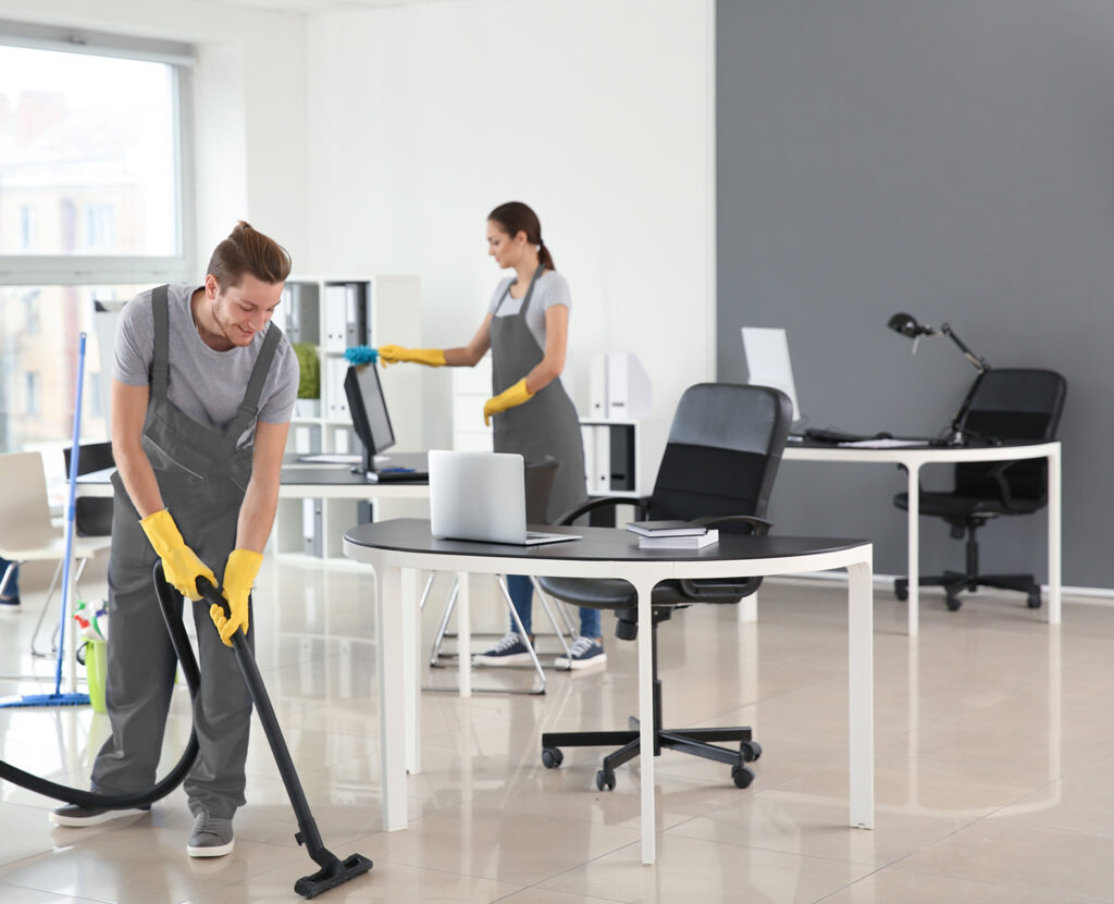 Two cleaners in uniforms vacuum and dust a modern office with laptops on desks.