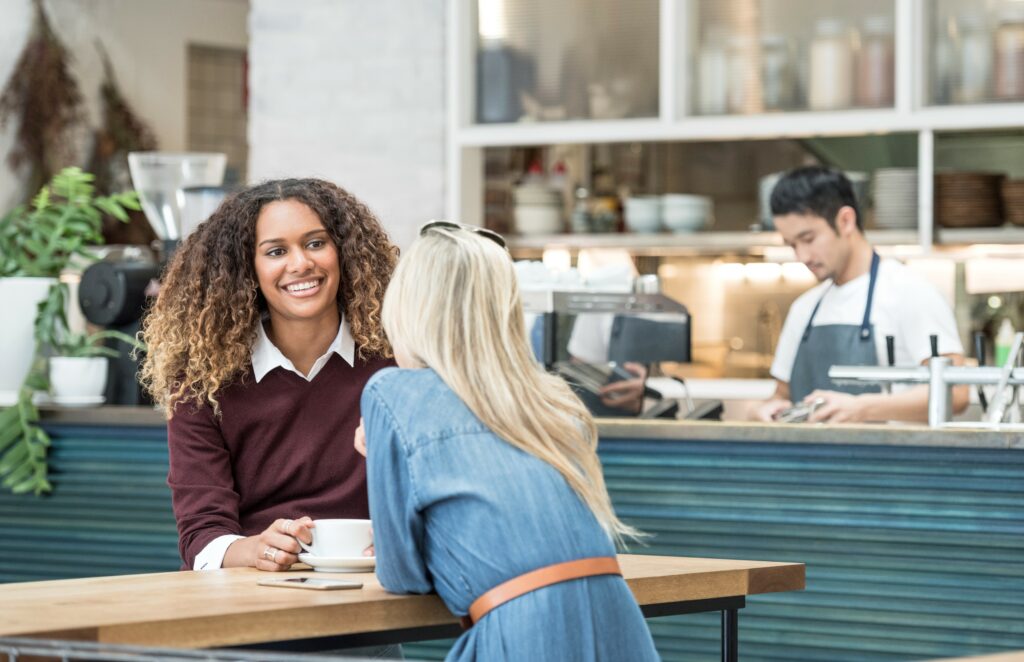 Two women sitting at a café table, smiling, with a barista preparing drinks in the background.