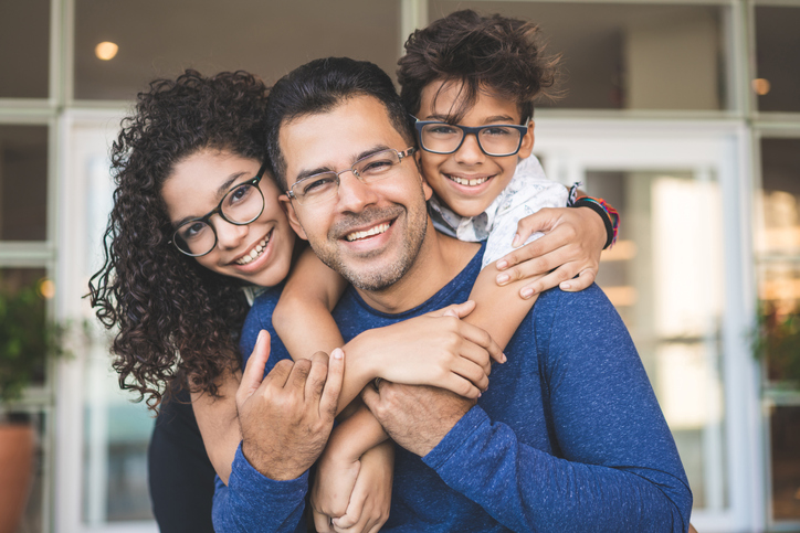 Smiling father with two children hugging him from behind, all wearing glasses, standing in front of a building entrance.