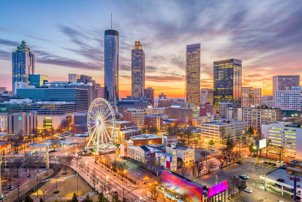 Skyline of a city at sunset with a prominent ferris wheel and illuminated skyscrapers.