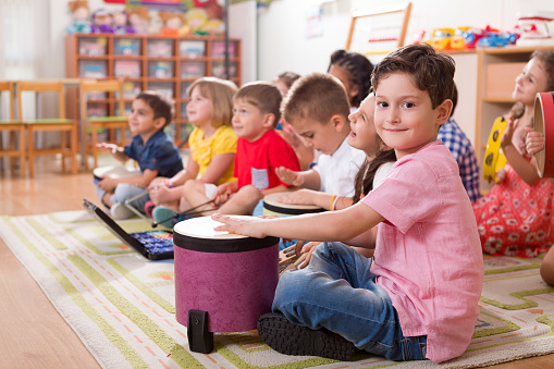 Children sitting on a rug playing hand drums in a classroom setting.