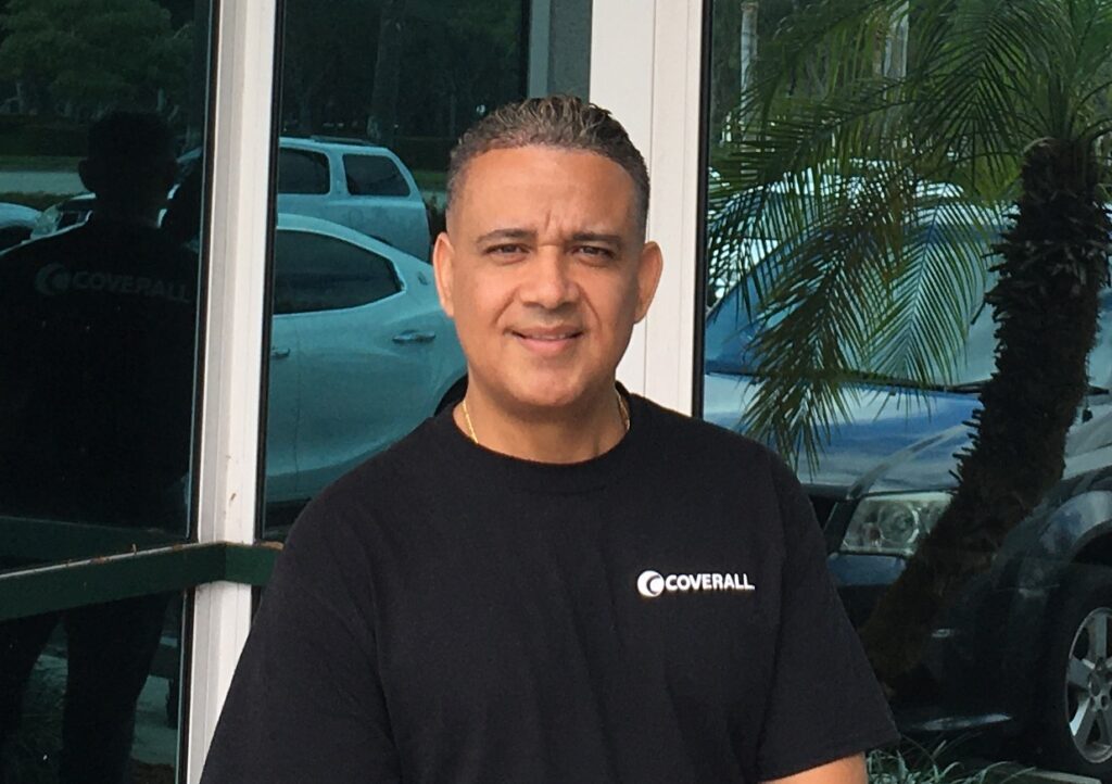 Man in a black "Coverall" shirt stands smiling near a glass window, with cars and palm trees in the background.