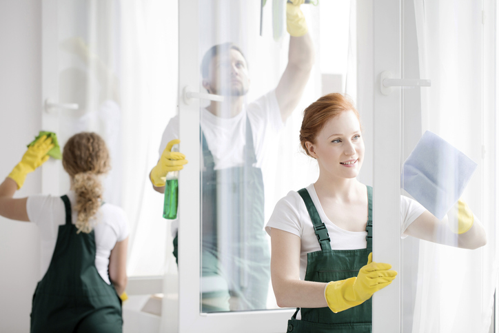 Two people in uniforms and gloves cleaning windows with spray bottles and cloths in a bright room.