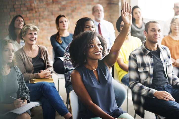 Audience in a seminar room, mainly seated with one smiling woman raising her hand to ask a question.
