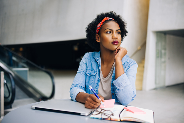 Woman in a denim shirt, sitting at a table, looks thoughtful while holding a pen. A laptop and notebook are in front of her.