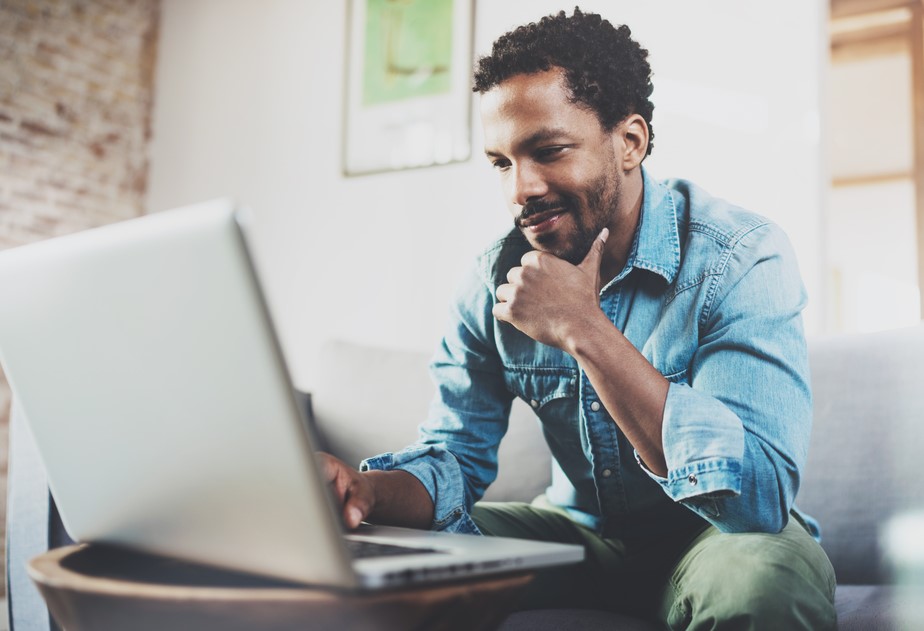 Person in denim shirt sitting on couch, using a laptop and looking thoughtful.