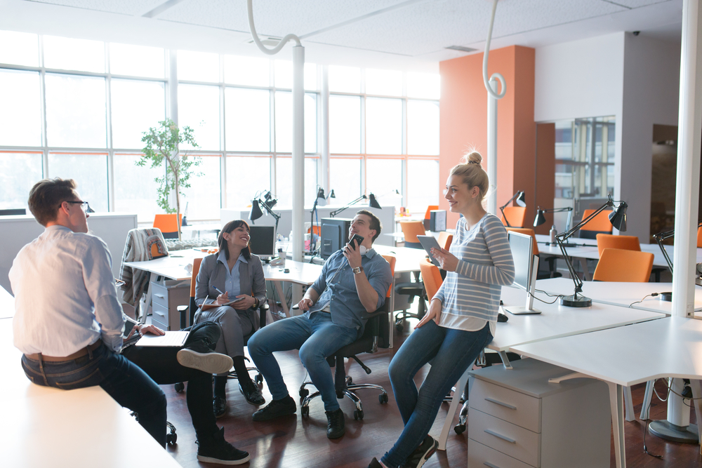 Colleagues gather and chat in a modern, bright office space with desks and large windows.