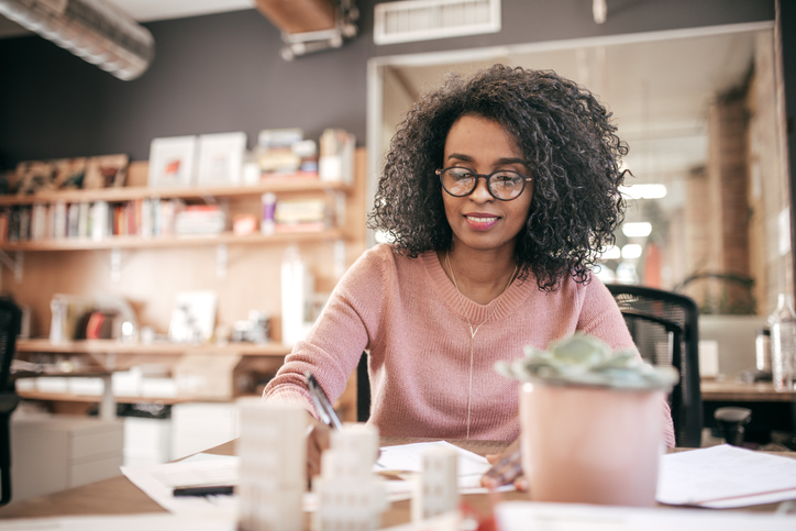 Woman in glasses working at a desk, writing on papers in an office setting with shelves and plant in background.
