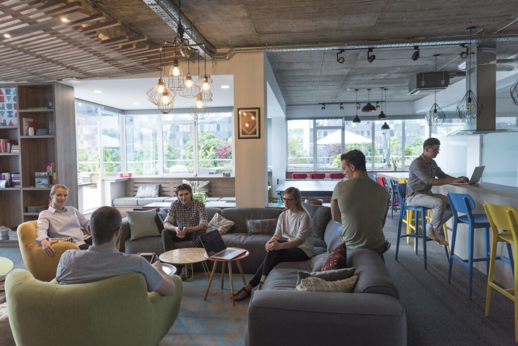 Modern office lounge with six people working on laptops and tablets, bright natural lighting, and colorful seating.
