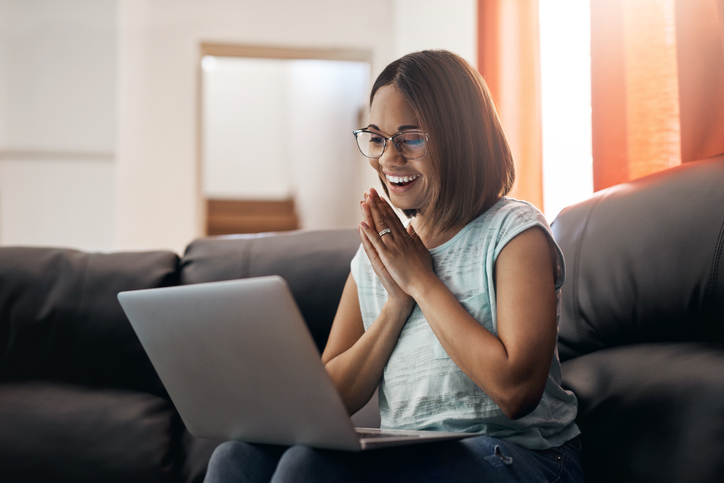 A woman sitting on a couch, smiling and using a laptop, with hands clasped together in excitement.