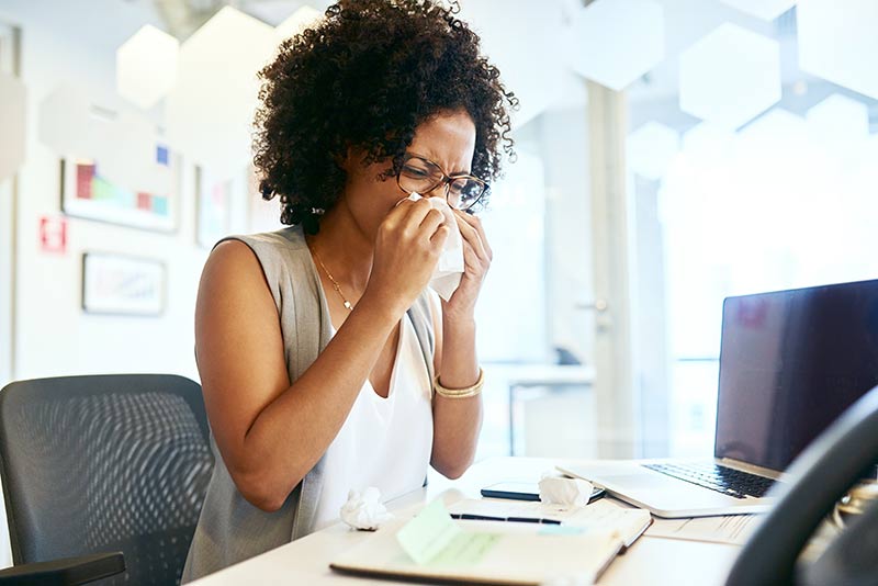 Person sneezing at a desk, using a tissue, with a laptop and notebook nearby in an office setting.