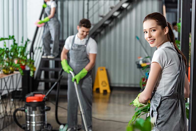 Two workers clean an indoor plant area, smiling and wearing gloves and overalls, with cleaning equipment visible.