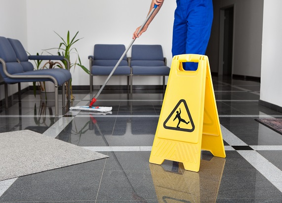 Person mopping a tiled floor beside a yellow caution sign in a lobby with chairs and a plant.