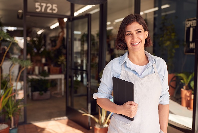 A smiling person holding a tablet stands in front of a plant shop with glass doors, wearing a light apron and shirt.