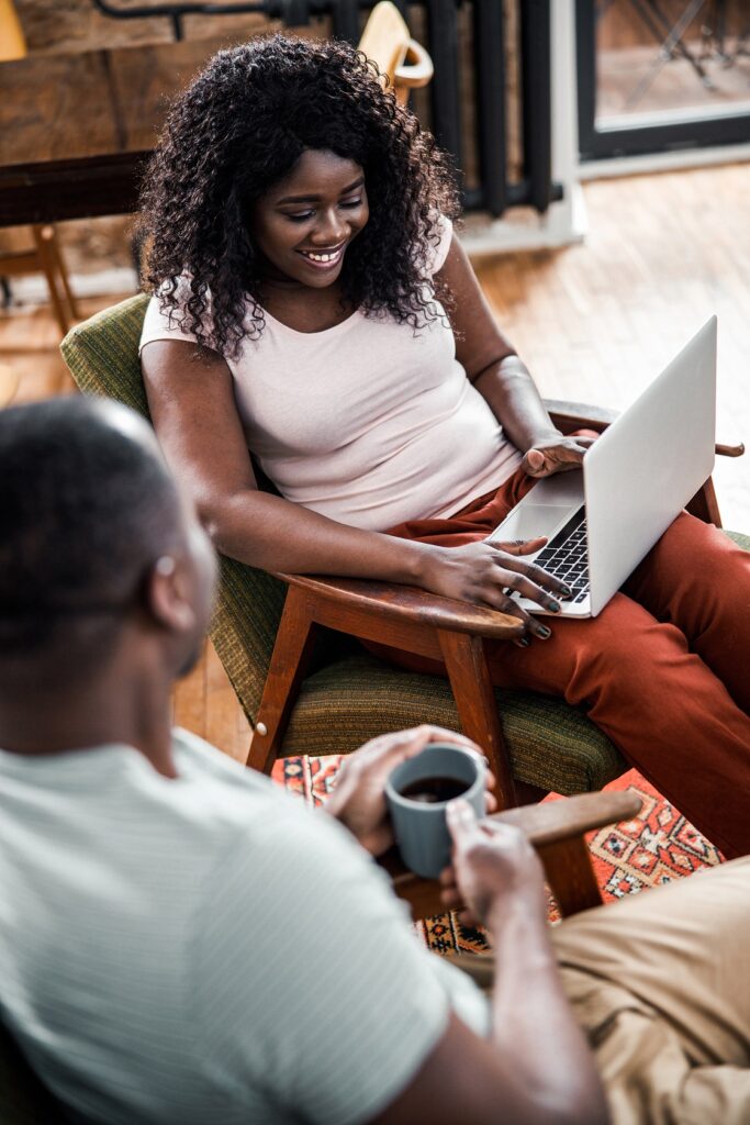 Two people sitting in a cozy room, one using a laptop and the other holding a coffee mug, smiling and conversing.