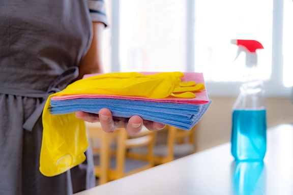 Person holds cleaning cloths and yellow gloves, with a spray bottle of blue liquid in the background.