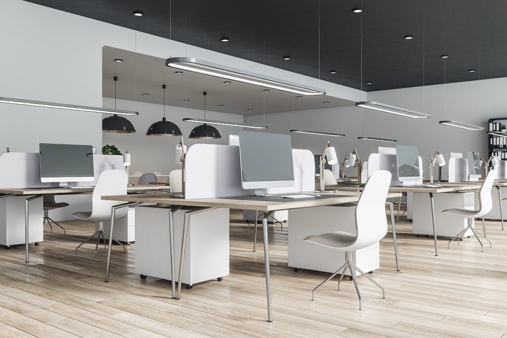 Modern office with white desks, computers, and chairs arranged in an open layout with overhead lighting.