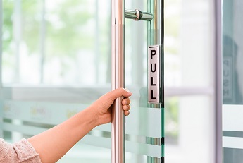 Hand pulling on a glass door handle with "PULL" sign, in a bright office environment.