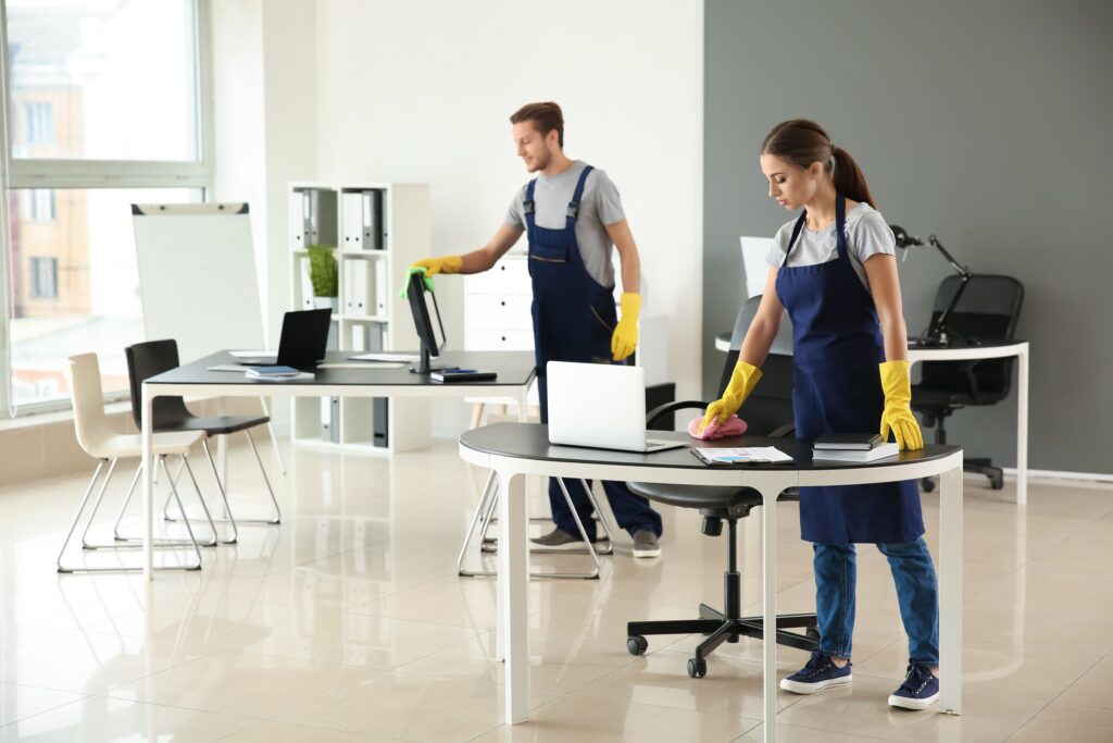 Two people in uniforms cleaning an office space, wiping tables with cloths and wearing yellow gloves.