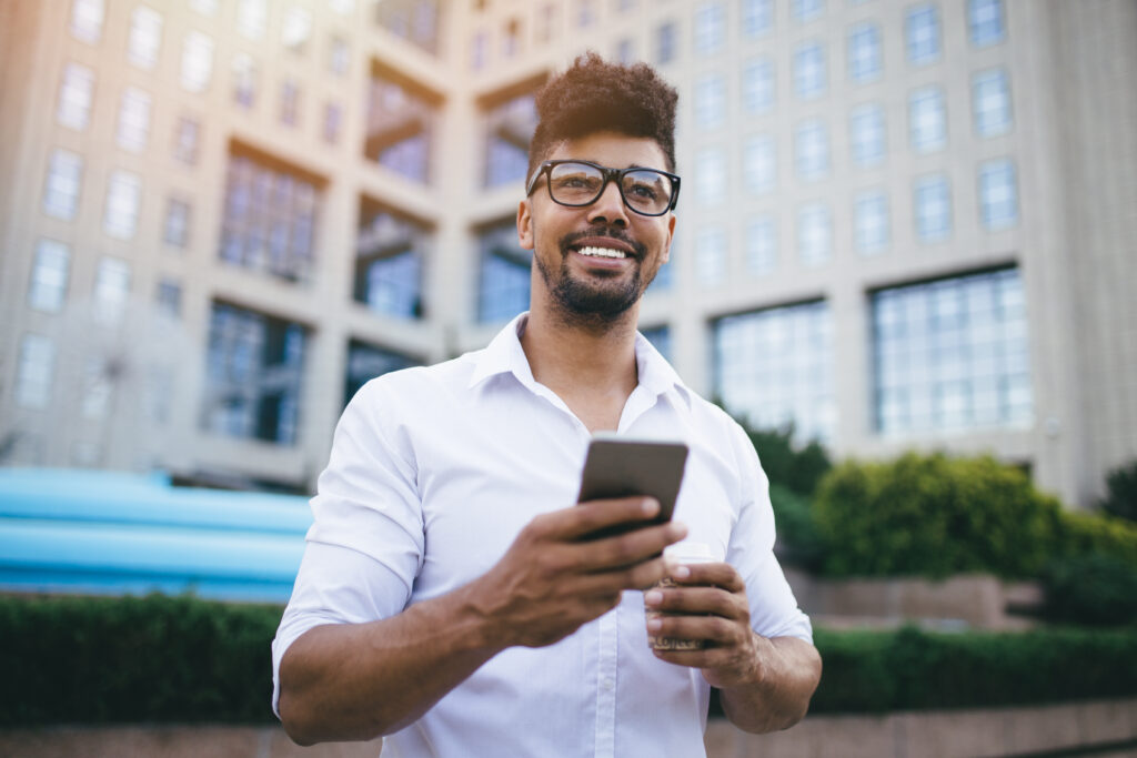 Man in glasses holding a phone and a coffee cup, smiling outdoors in front of a modern building.