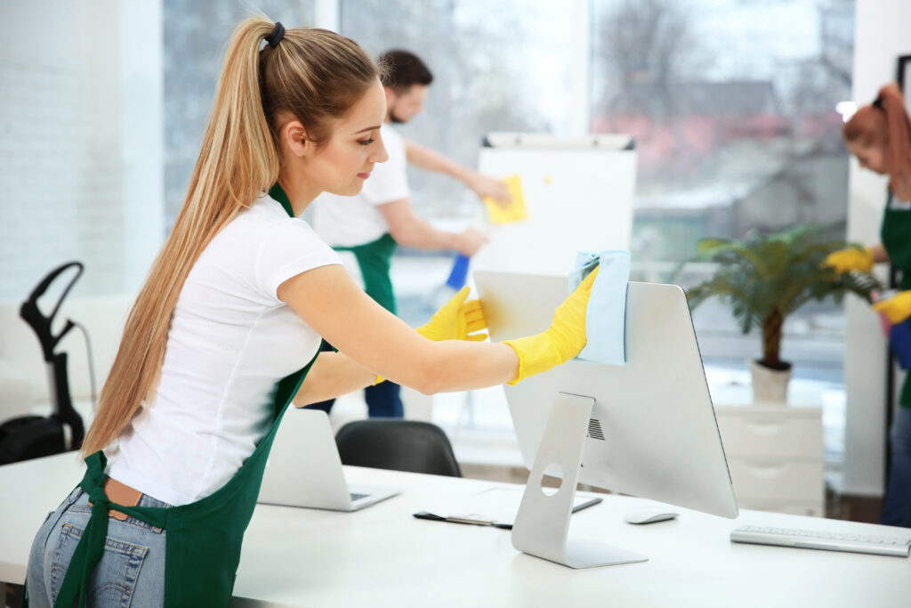 Person cleaning a computer monitor in an office, wearing yellow gloves and a green apron.