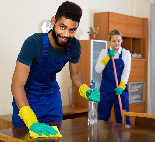 Two cleaners in blue uniforms clean a room, one wiping a table and the other mopping the floor.