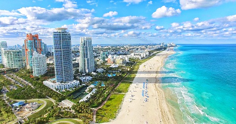 Aerial view of a coastal city with skyscrapers, a sandy beach, and turquoise ocean under a partly cloudy sky.