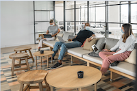 Three people in masks use laptops in a modern lounge area with round tables, glass walls, and patterned flooring.