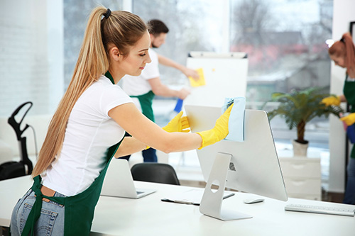Office cleaning staff wiping desks and monitors, wearing aprons and gloves, in a bright workspace.