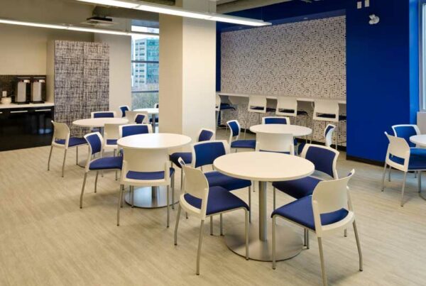 Modern breakroom with round tables, blue chairs, and a counter with coffee machines, featuring bright lighting and large wind