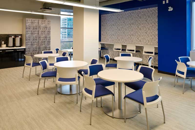Modern breakroom with round tables, blue chairs, and a counter with coffee machines, featuring bright lighting and large wind