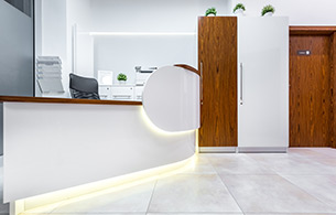 Modern office reception area with white desk, wood accents, plants, and a closed door on the right.