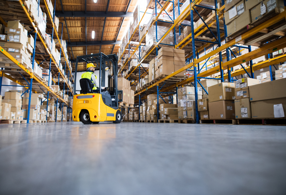 Worker operating a yellow forklift in a warehouse aisle with stacked boxes on shelves.