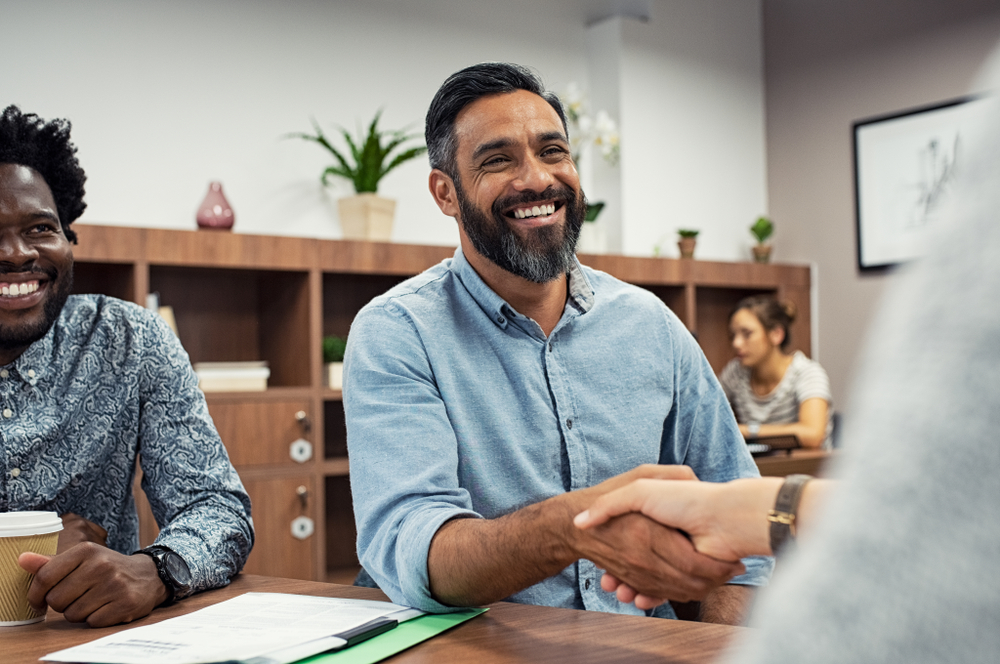 Smiling man in office shakes hands with unseen person, colleague smiles beside him. Shelves and plants in the background.