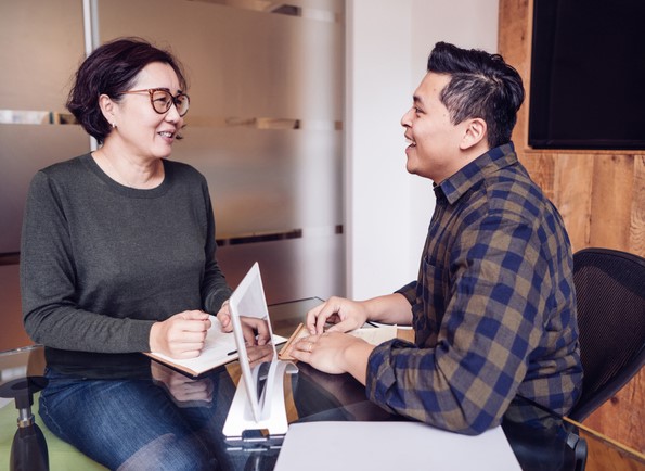 Two people sitting at a desk, having a conversation with a laptop open.