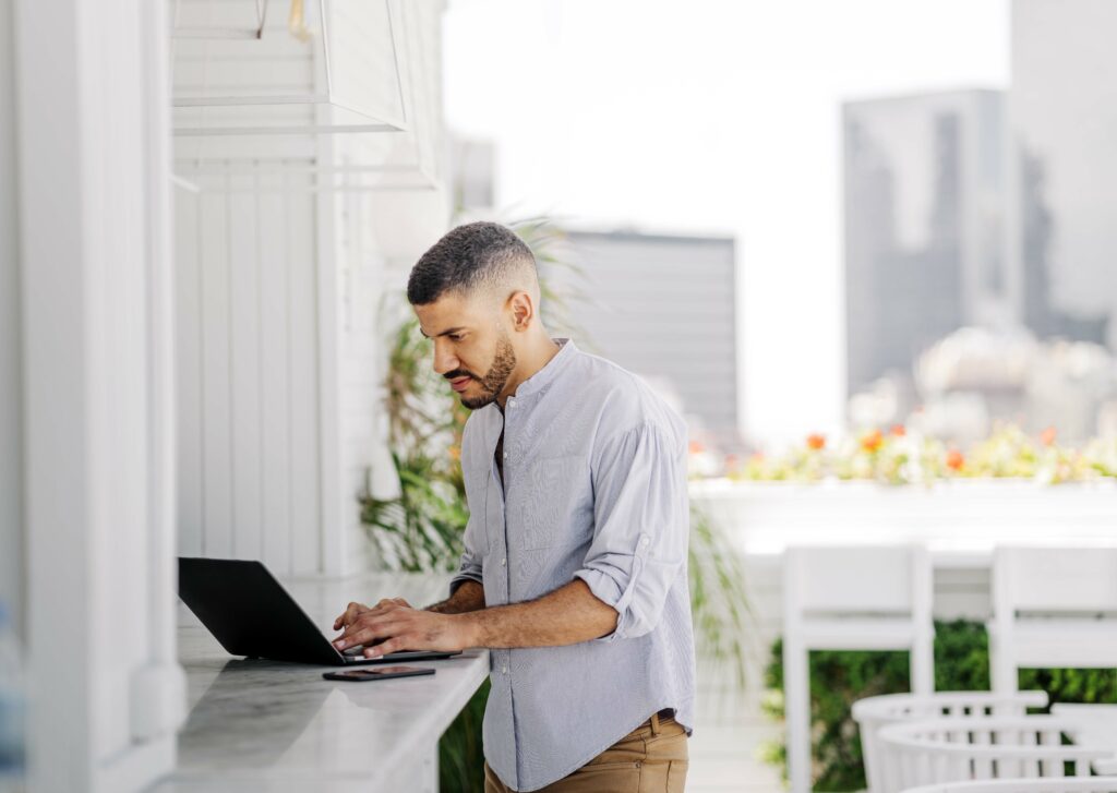Man working on laptop outdoors, standing at counter overlooking city view.