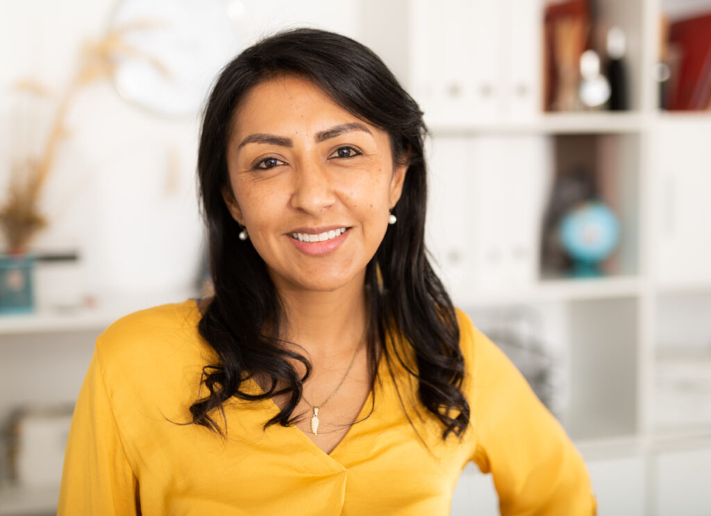 Smiling woman with long dark hair in a yellow top stands in an office with shelves in the background.