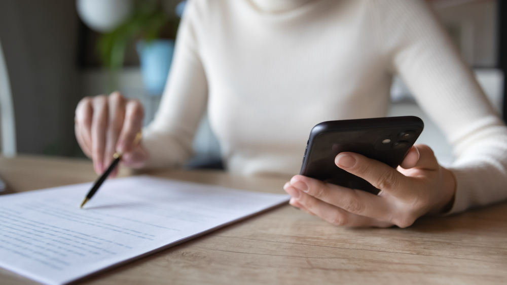 Person reviewing document while holding a smartphone, focused on multitasking.