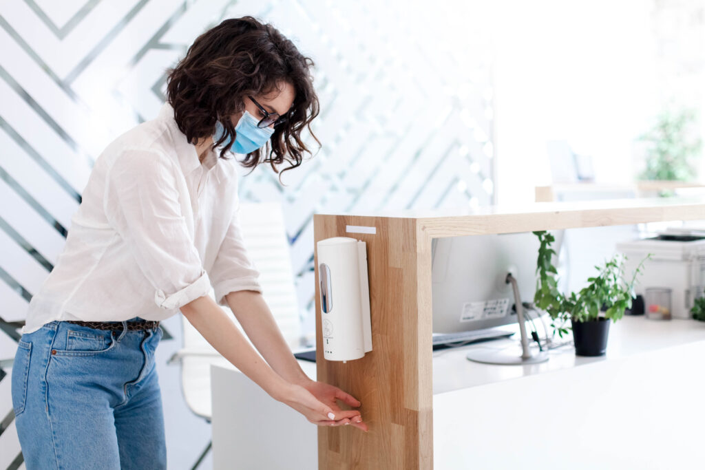 Person in mask sanitizing hands at a modern reception desk with a computer and plants.