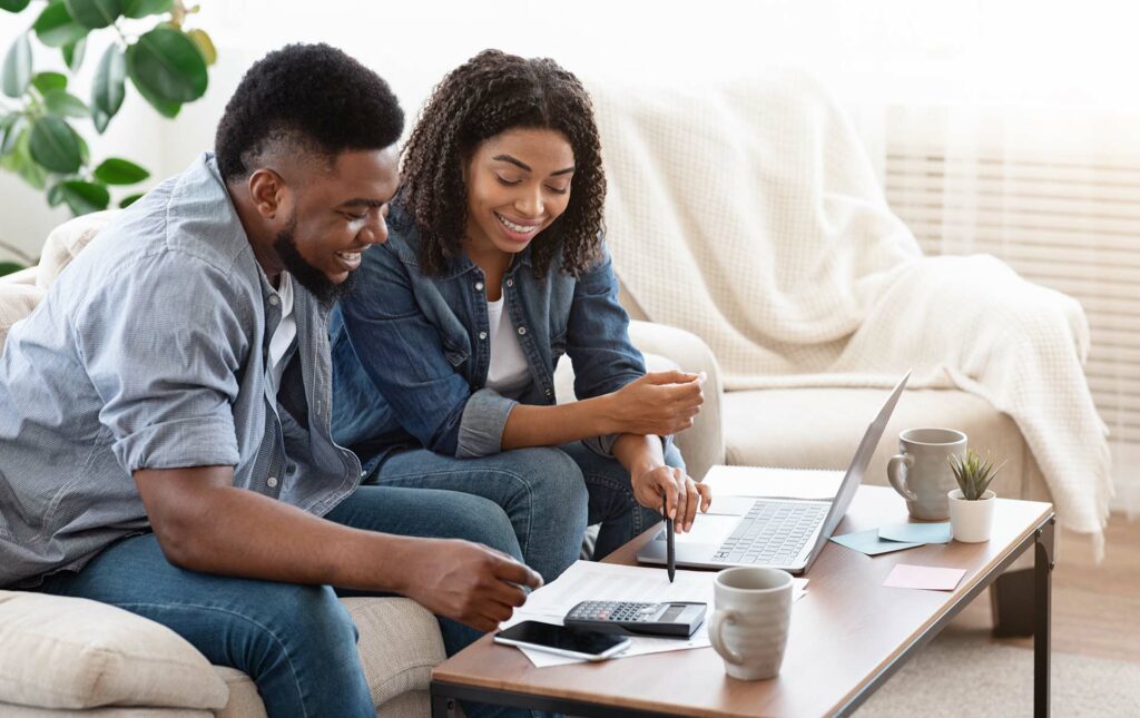 Couple smiling while budgeting together using a laptop, calculator, and papers on a table in a cozy living room.