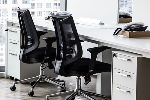 Empty office with two black ergonomic chairs and white desks in front of large windows.