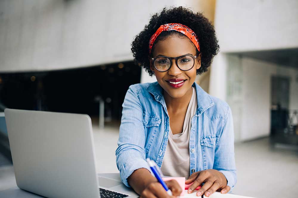 A woman with glasses and a red headband writes in a notebook, smiling, seated at a desk with a laptop.