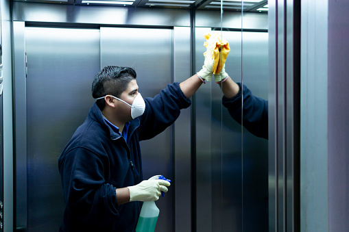 Person wearing a mask and gloves cleaning elevator mirrors with cloth and spray bottle.