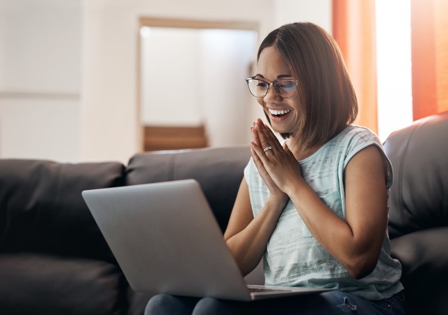 Person smiling and clasping hands, seated on a couch using a laptop, ambient indoor lighting.