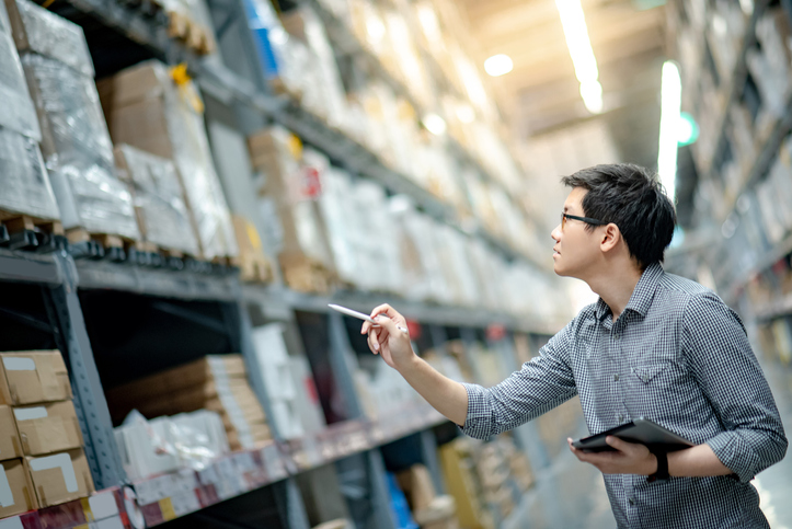 Person with tablet inspecting shelves in a warehouse aisle.